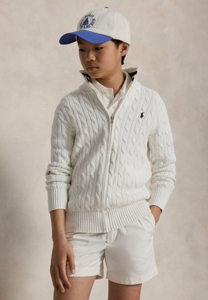 Un jeune homme porte un cardigan blanc en maille torsadée avec fermeture éclair, un short blanc et une casquette blanche à visière bleue, sur un fond neutre.