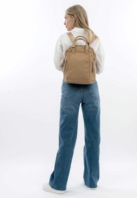 Zaino tan realizzato in tessuto resistente, dotato di due tasche laterali, maniglie superiori e spallacci regolabili. Texture liscia con design minimalista.