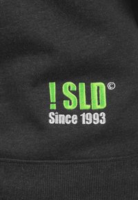 Zwarte sweatshirt met groene en witte geborduurde tekst: "! SLD" en "Sinds 1993." Zacht materiaal met geribbelde onderkant.