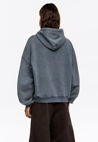 Oversizowa bluza z przetartym błękitnym materiałem, z dużym kapturem, opuszczonymi ramionami i prążkowanym wykończeniem na dole. Połączona z luźnymi, ciemnymi spodniami.