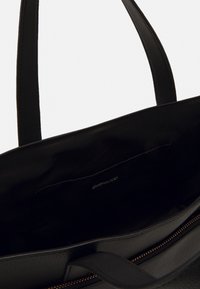 Bolso tote de cuero negro con dos asas, presenta una textura suave y una cremallera dorada. El interior está forrado con tela negra y una etiqueta con el logo.
