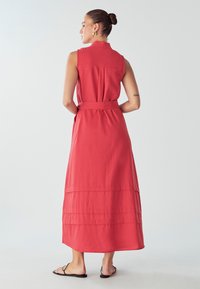 Červené maxi šaty bez rukávov s opaskom v páse, s vrstveným dizajnom a klasickým golierom. K šatám sú obuté ploché sandále. Hladký povrch látky.