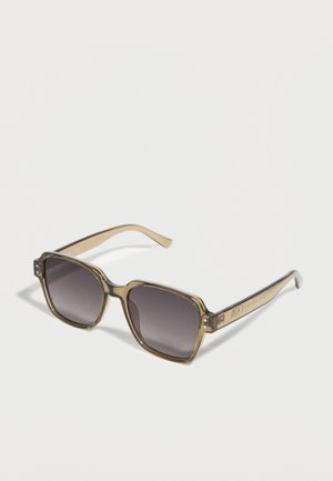 Quadratische Sonnenbrille mit durchscheinend beigen Bügeln und dunklen Verlaufsgläsern, auf einer weißen Oberfläche liegend.