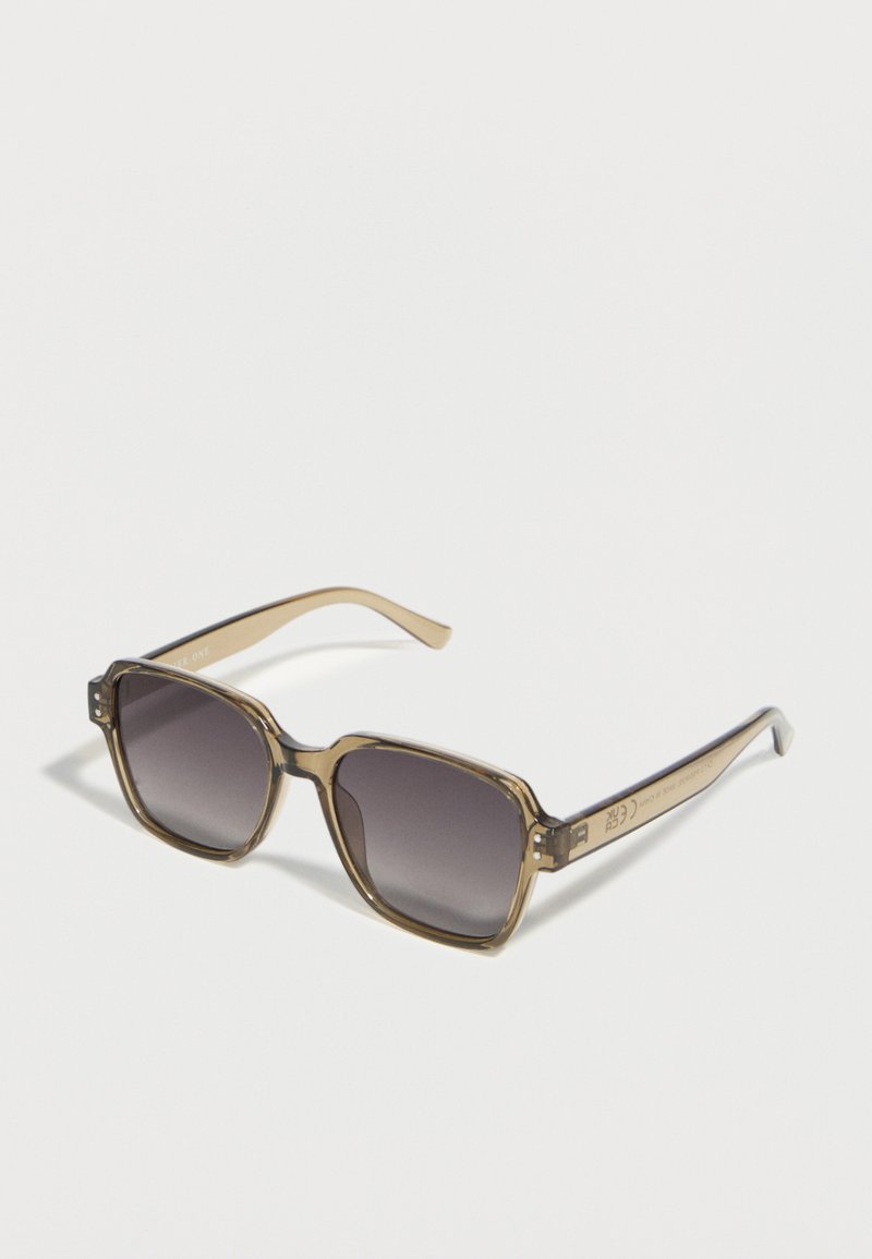 Γυαλιά ηλίου με τετράγωνο πλαίσιο, διάφανα μπεζ μπράτσα και σκουρόχρωμους φακούς με βαθμιαία απόχρωση, τοποθετημένα πάνω σε λευκή επιφάνεια.