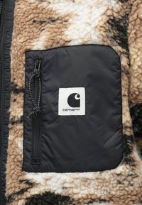 Chaqueta de forro polar en marrón y crema con un patrón texturizado. Cuenta con un bolsillo con cremallera negra y una etiqueta con el logo blanco de Carhartt.