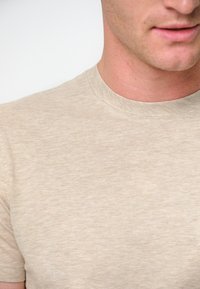 Beige bomullströja med rundad hals, slät textur, korta ärmar och ett subtilt, melerat mönster. Nærbild.