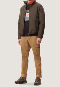 Olivengrøn letvægtsjakke, sort sweatshirt med grafik, tan cargo-bukser og robuste sorte vandresko. Lynlås- og patch-detaljer synlige.