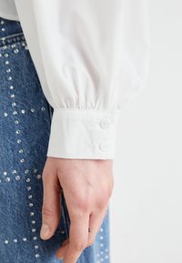 Chemise blanche à manches longues avec des poignets froncés, dotée d'une fermeture à double bouton. La main repose sur un jean bleu avec des détails en clous argentés.