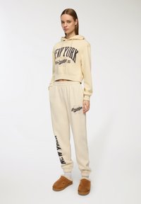 Kremowa, krótka bluza z kapturem z dużym nadrukiem "NEW YORK" oraz spodnie dresowe z elastycznym paskiem i podobnym nadrukiem. Brązowe, puszyste kapcie.