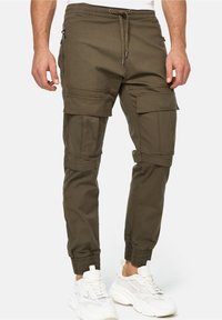 Olivengrøn cargo-bukser med lynlåslommer, tætsiddende pasform, justerbar snøre i taljen og elastiske manchetter, parret med hvide chunky sneakers.