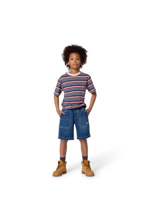 Enfant aux cheveux bouclés portant un t-shirt rayé, un short en denim et des bottes marron, debout les mains dans les poches sur fond blanc.