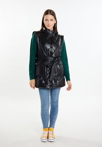Gilet smanicato imbottito nero con colletto alto, dettaglio con cintura e due tasche frontali. Indossato sopra un maglione in maglia verde e jeans di denim.