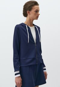 Felpa navy con zip e cordini bianchi, caratterizzata da una vestibilità rilassata e polsini a righe, realizzata in un tessuto liscio e morbido. Abbinata a pantaloni corti.