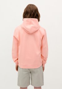 Maglione con cappuccio rosa realizzato in tessuto morbido, con una vestibilità comoda, polsini a coste e tasca a marsupio. Indossato con pantaloni corti grigi chiari.