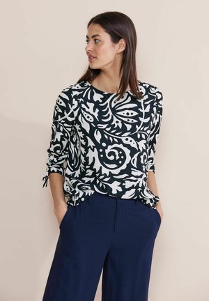 Frau mit glatten, dunklen Haaren, die ein langärmliges Oberteil mit schwarz-weißen Blumenmuster und marineblauen Hosen trägt, die Hände in den Taschen, nach links schauend.