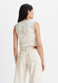 Cropped denim vest in pale beige with blue tie-dye patterns, featuring a simple design and a back pocket detail. Worn with high-waisted jeans.