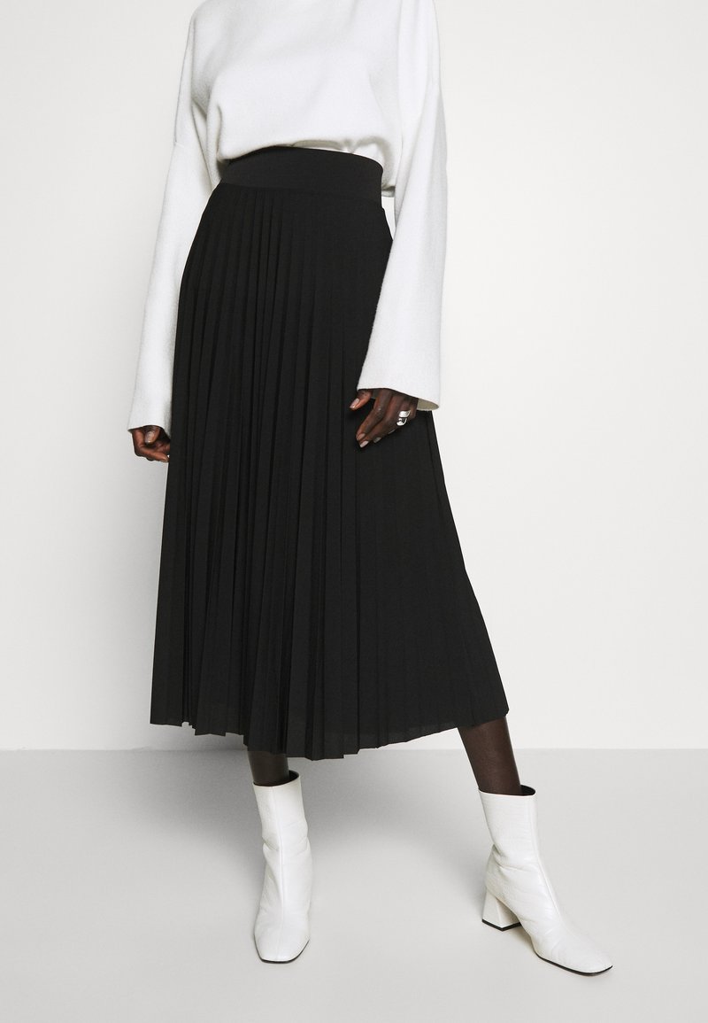 Black pleated skirt with an elastic waistband, paired with a white long-sleeve top and white ankle boots. Smooth fabric texture.