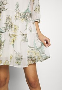 Robe mini florale en tissu léger et transparent. Présente des motifs floraux jaunes et teal, un design à volants et une silhouette fluide.