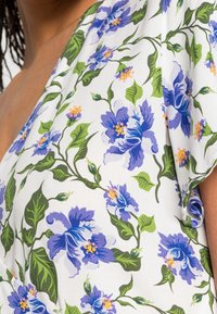 Haut portefeuille fleuri avec un fond blanc orné de grandes fleurs violettes et de feuilles vertes. Tissu doux avec un design de manches drapées.