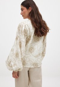 Valkoinen paita, jossa on monimutkainen vaaleanbeige kukka-embroidery ja puffihihat. Paita on rento malli ja valmistettu kevyestä, tekstuurillisesta materiaalista.