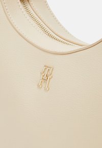 Beżowa skórzana torba o teksturowanej powierzchni, z akcentem w postaci złotego logo i zamknięciem na zamek wzdłuż górnej krawędzi.