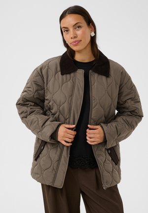 Femme aux longs cheveux bruns portant une veste matelassée taupe avec col marron foncé, chemise noire, pantalon marron et boucles d’oreilles argentées, de face.