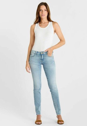 Femme debout, portant un haut blanc sans manches, un jean bleu clair et des sandales ouvertes marron, sur un fond clair uni.