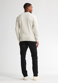 Pulover bej deschis cu maneci lungi, cu tivuri ribbate, purtat cu pantaloni negri skinny și adidași albi. Vedere din spate, fundal simplu.