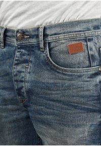 Jeans de mezclilla azul con un lavado desgastado, que presentan un parche de cuero etiquetado como "BLEND", un botón de metal y costuras amarillas alrededor de los bolsillos.