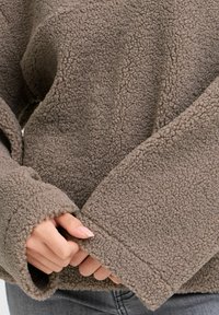 Brun fleece-pullover med et tekstureret, hyggeligt stof og en løs pasform. Ærmerne er brede og ender i syede manchetter.