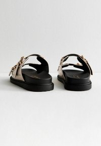 Sandalias de plataforma negras con tiras de cuero sintético beige, que presentan un detalle de lazo y un diseño de espalda abierta. Plantilla texturizada y acabado suave.