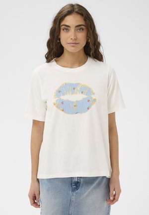 Młoda kobieta z kręconymi włosami, ubrana w biały T-shirt z dużym nadrukiem ust w niebiesko-żółte paski i małymi kolorowymi kropkami, w połączeniu z dżinsową spódnicą.