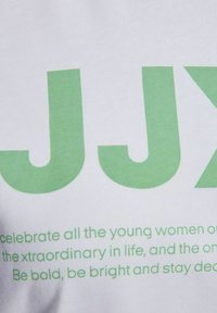 Camisa de algodón blanca con texto grande en verde "JJX" y texto más pequeño debajo en verde, con una textura suave y un diseño casual.