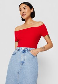 Top rosso a spalle scoperte realizzato in tessuto a coste, abbinato a una gonna di jeans azzurro chiaro con spacco frontale e tasche anteriori.