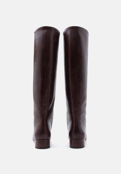 Ζευγάρι ψηλές, σκούρες καφέ δερμάτινες μπότες με χαμηλά τακούνια, εμφανιζόμενες από πίσω σε ουδέτερο φόντο.