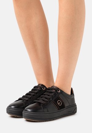 Pieds portant des baskets en cuir noir à lacets, avec un logo rond sur le côté et des semelles épaisses en caoutchouc, sur un fond uni.