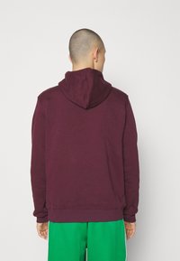 Mörkröd hoodie med snöre i huvan, långa ärmar och ribbade muddar. Bärs över gröna shorts, vilket visar en jämn tygtextur.