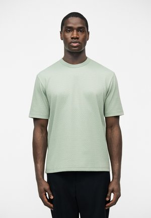 Homme debout, portant une chemise à manches courtes vert clair avec un motif subtil et un pantalon noir, sur un fond blanc uni.