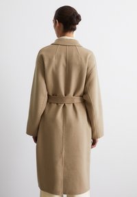 Cappotto trench in lana beige con cintura, maniche lunghe e vestibilità rilassata. Presenta una fessura sul retro e cuciture pulite per un dettaglio minimale.