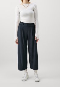 Bílý top s dlouhým rukávem spárovaný s námořnickými širokými kalhotami s pruhy, které mají dvojité knoflíky v pase. Model má na sobě bílé tenisky a ponožky.