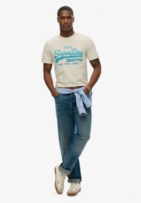 Camiseta de algodón beige con gráfico en turquesa "SuperDry", combinada con vaqueros de mezclilla azules y zapatillas crema con suelas marrones. Camisa azul claro atada a la cintura.