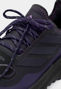 Gros plan sur une chaussure de sport violet foncé avec des lacets rayés réfléchissants, des détails de couture en zigzag et une semelle en caoutchouc à grip texturé.