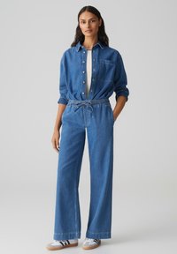 Chemise en denim avec des boutons sur le devant et une poche poitrine, associée à un pantalon en denim à jambes larges avec une taille élastique et un cordon de serrage, le tout en bleu clair.