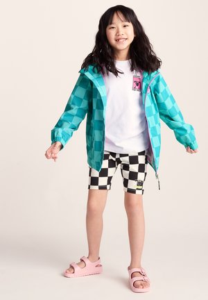 Young girl smiling, wearing turquoise checkered jacket, white t-shirt, black and white checkered shorts, and pink sandals, standing with arms slightly out.