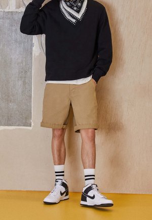 Persoon draagt een zwart shirt met lange mouwen, een zwart-wit geblokte bandana, beige opgerolde korte broek, witte sokken met zwarte strepen en witte Nike-sneakers.