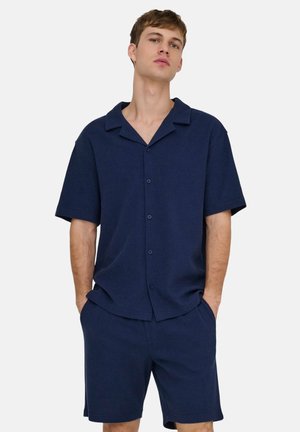 Jeune homme portant une chemise bleu marine à manches courtes boutonnée et un short assorti, les mains dans les poches, sur un fond clair uni.