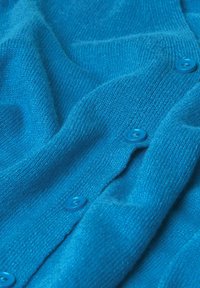 Türkise Strickjacke aus strukturiertem Stoff, mit runden blauen Knöpfen entlang der Vorderseite, die ein weiches, gemütliches Erscheinungsbild zeigt.