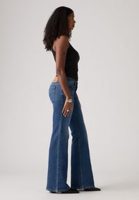 Jeans en denim bleu évasés avec une taille haute, dotés d'une étiquette en cuir à l'arrière. Portés avec un débardeur noir et des chaussures à talons.