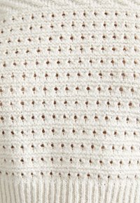Strukturierter Strickstoff in Creme mit kleinen braunen Punkten. Verfügt über einen gerippten Saum am Boden. Das Muster hat gleichmäßig verteilte Löcher.