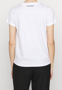 Valkoinen puuvillapaita pyöreällä kaula-aukolla ja lyhyillä hihoilla, jossa on musta "KARL LAGERFELD" -logo yläselässä, minimalistinen muotoilu.